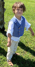 Load image into Gallery viewer, Little boy with a cute grin wearing the double breasted waistcoat with wood buttons, white linen button down shirt with the sleeves rolled up and buttoned with the sleeve tabs and natural linen pants
