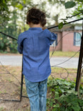 Load image into Gallery viewer, little boy showing the back of the blue chambray button down with the pleat detail and curved hem