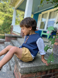 Load image into Gallery viewer, Little boy sitting on a step wearing the off white linen button back shirt, textured chambray vest in blue and pleated cuffed shorts in khaki
