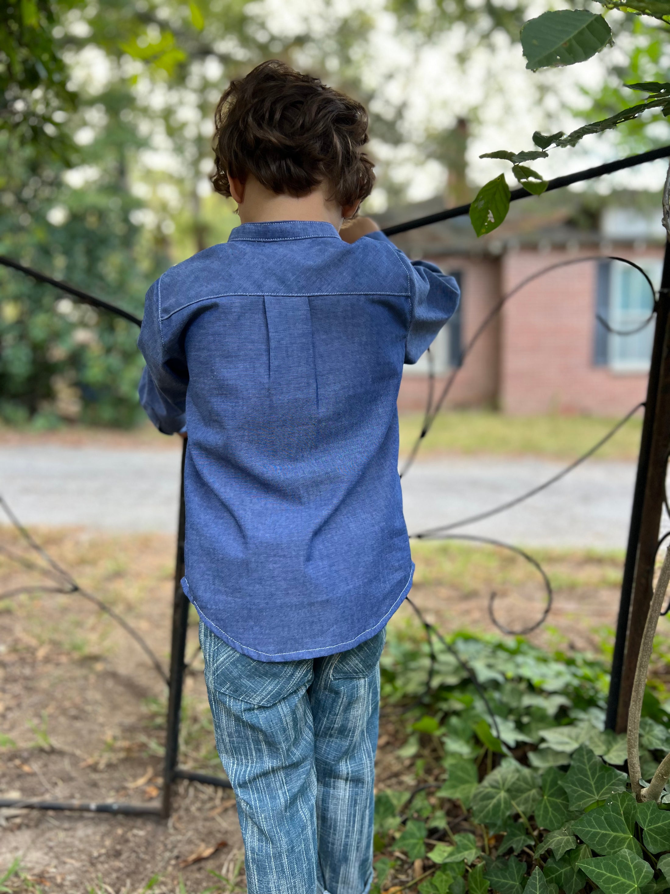 little boy showing the back of the blue chambray button down with the pleat detail and curved hem