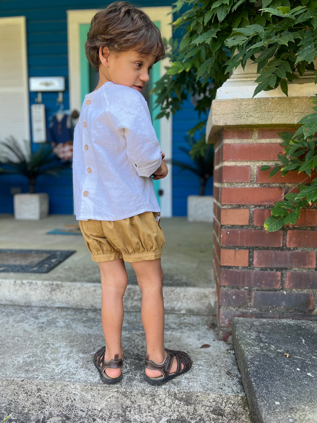 Little boy wearing the button back off white linen shirt and pleated cuff shorts.  He has his back to the camera to show he buttons on the shirt and the elastic waist on the back of the shorts