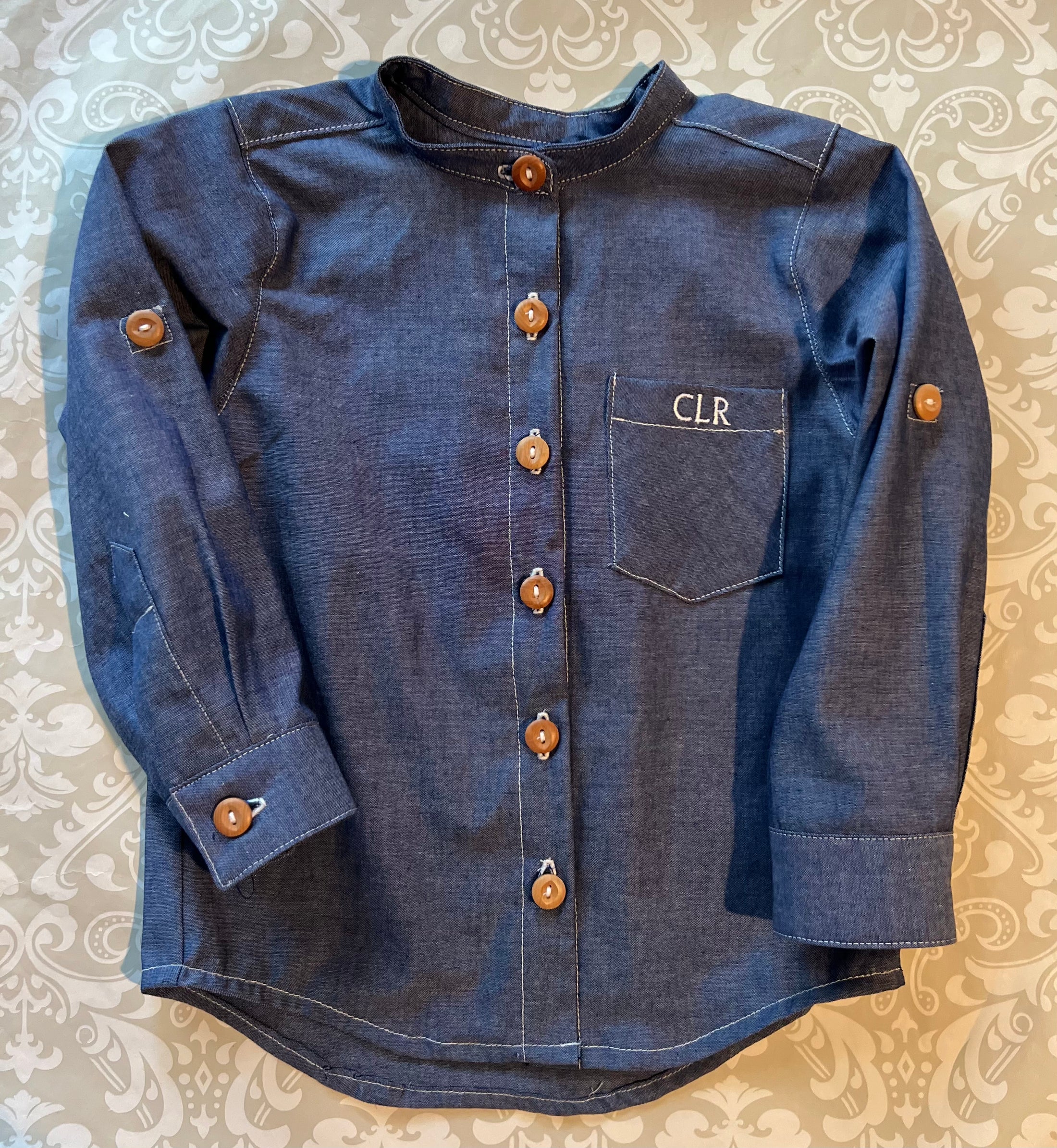 a flat view of the blue chambray button down shirt showing all the detail of the white contrast stitching, wood buttons and pocket monogram