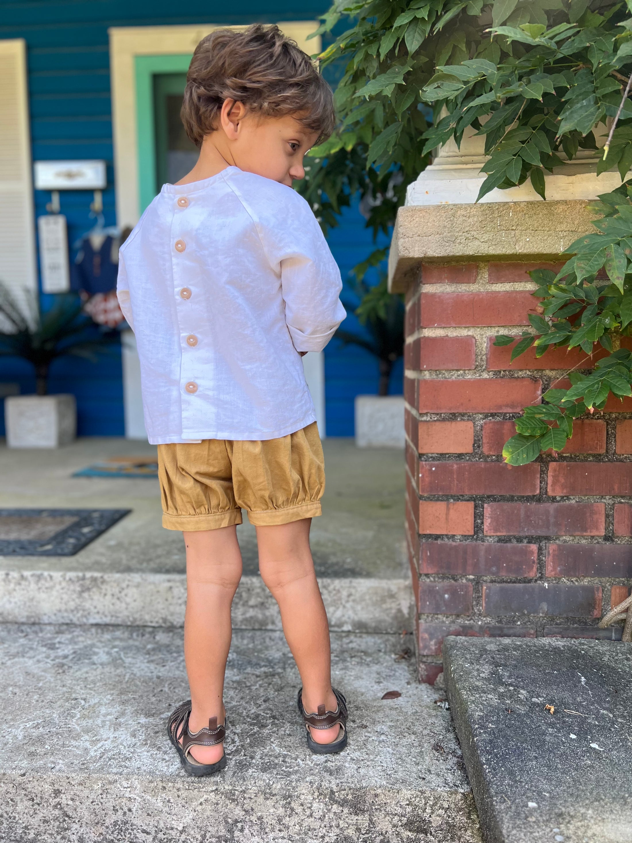 little boy standing with his back to the camera wearing the off white linen button back shirt and pleated cuffed shorts in Khaki color