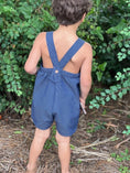 Load image into Gallery viewer, little boy with his back to the camera showing the detail of the straps and wooden button on the blue textured chambray vintage style romper 

