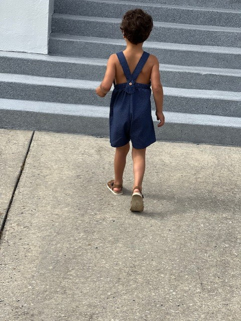 Little boy walking with his back to the camera showing the detail of the straps and shorts style of the romper