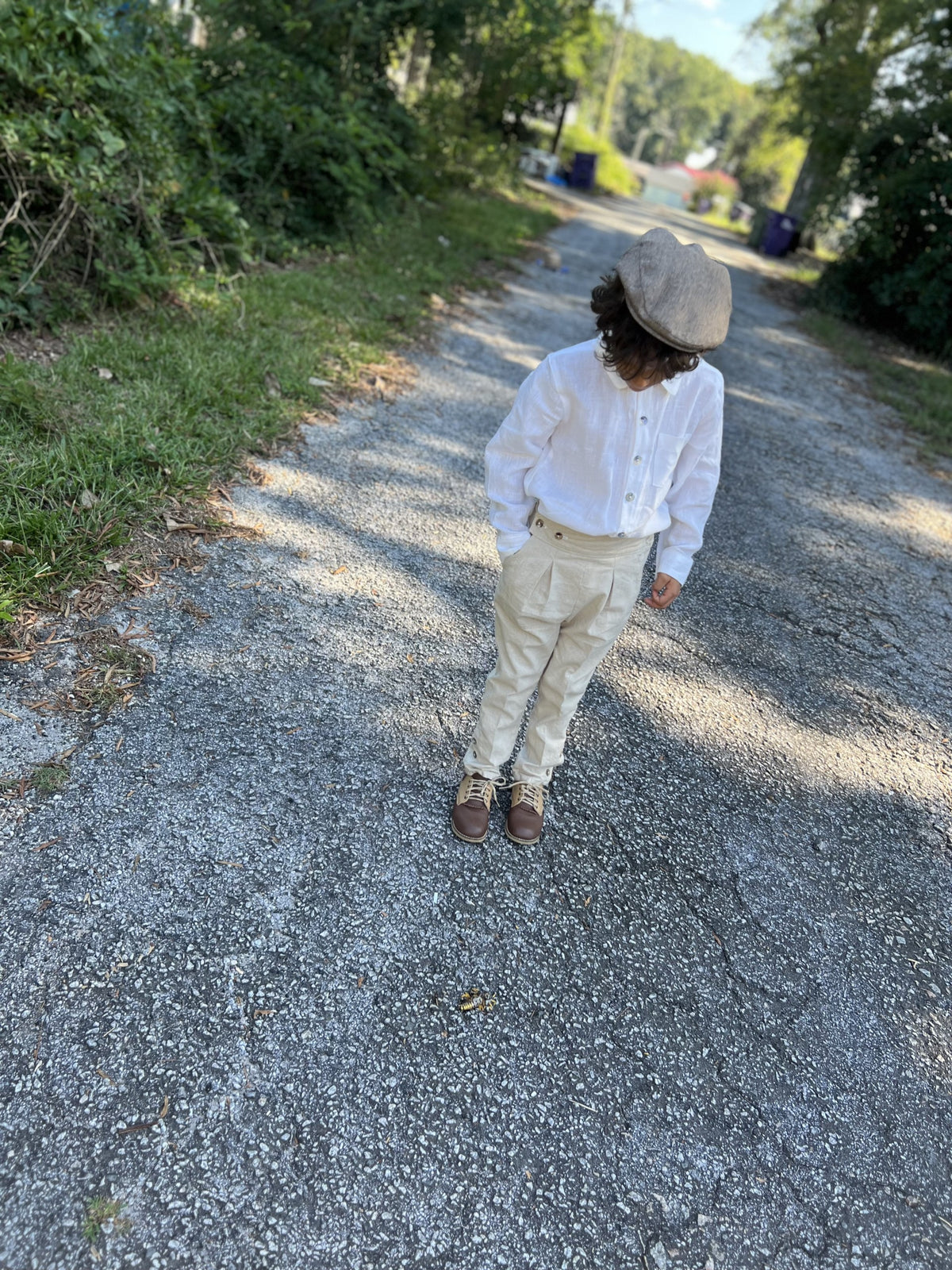 A child standing on a gravel path outdoors, wearing a white long-sleeve button-up shirt, beige trousers, brown shoes, and a taupe flat cap.