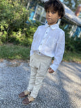 Cargar la imagen en la vista de la galería, A young boy wearing a white long-sleeve button-up shirt, beige trousers, and brown shoes, standing outdoors on a gravel path with green foliage and a house in the background.
