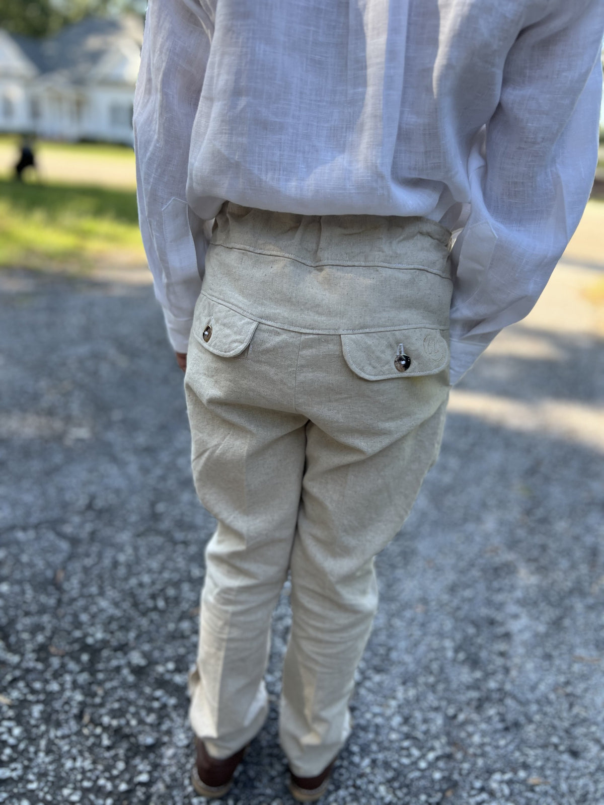 A person viewed from behind wearing light beige linen trousers with buttoned flap back pockets and a white linen shirt, standing on a gravel path outdoors.