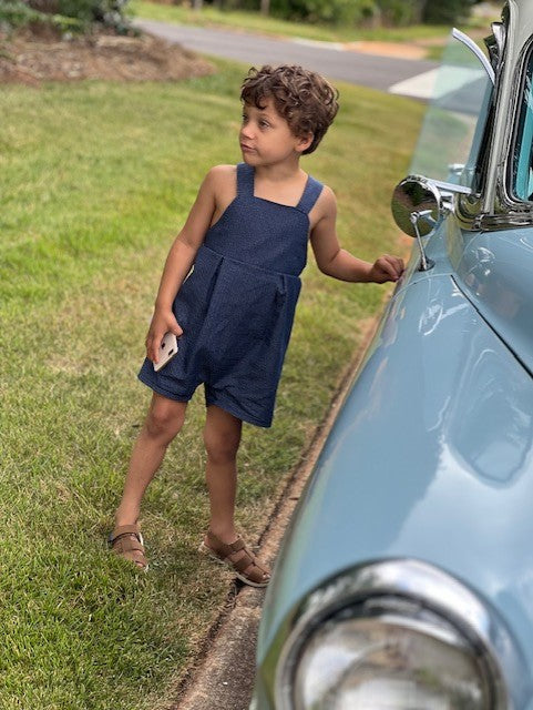 Little boy standing beside an old car.  He is wearing the vintage style romper in blue textured chambray