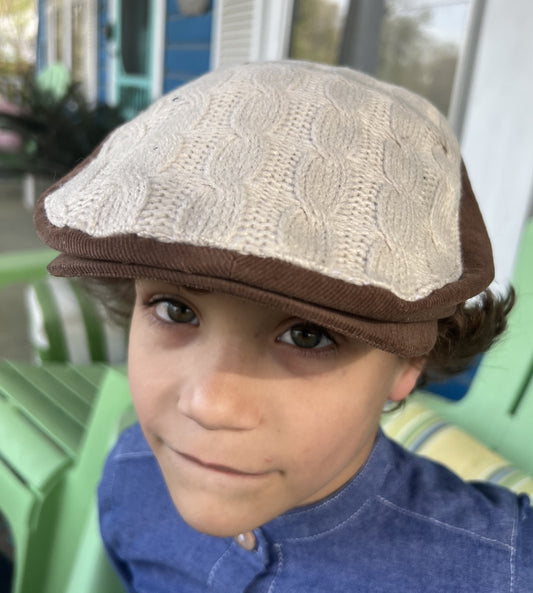 boy wearing a brown and cream newsboy cap.  the top is a cream cable knit sweater and the brown is a corduroy.